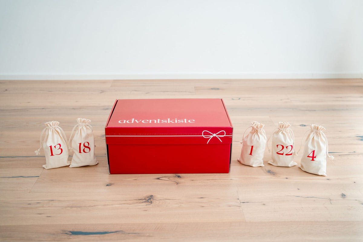 Geschlossene rote Adventkiste, daneben fünf cremefarbene Stoffsäckchen mit roten Zahlen.